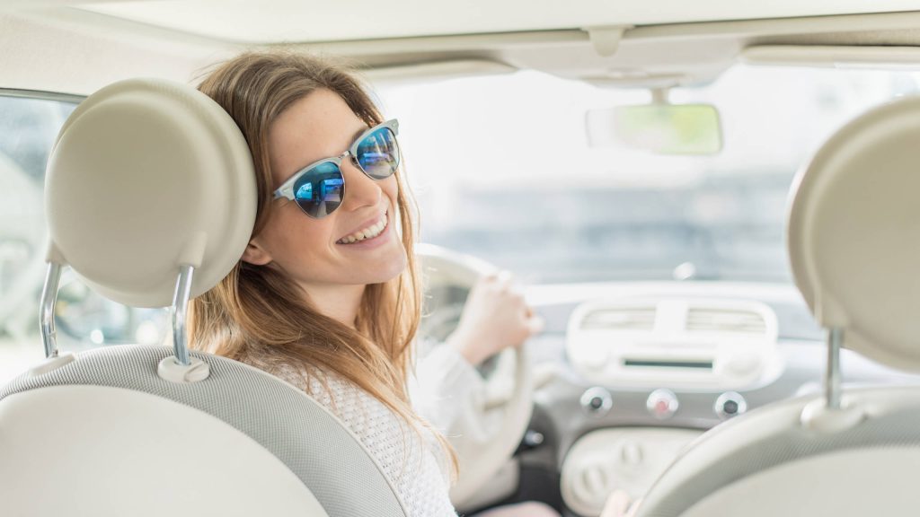 A woman wearing sunglasses sits in the driver's seat and smiles over her should towards the backseat; she just saved money by refinancing an auto loan