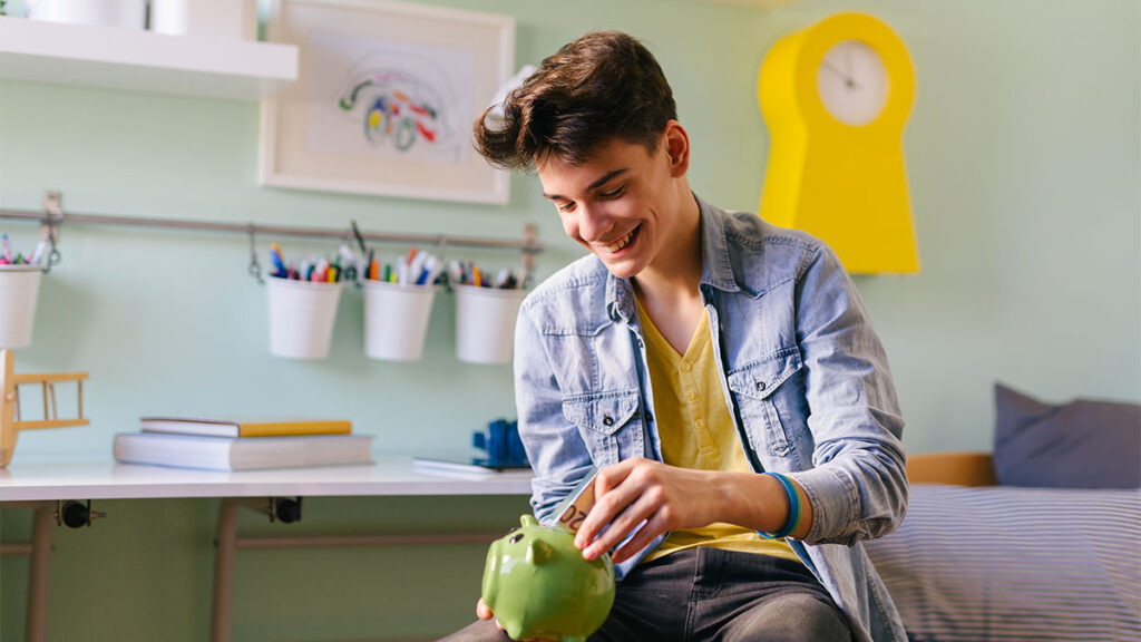 Teenage boy sitting in room putting coins into a piggy bank for a blog about budgeting tips for teens